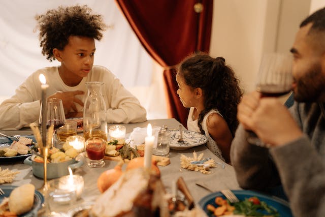 A young girl and boy talking at the Thanksgiving table while an adult watches on, smiling. 
